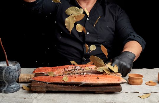 adult man in a black shirt pours white coarse salt and a dry bay leaf on a fresh salmon filet, cooking process, dark background