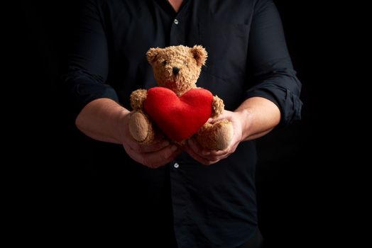 adult man in a black shirt holds a brown teddy bear with a red heart on a dark background,  concept of congratulations on the holiday of Valentine's Day on February 14, low key