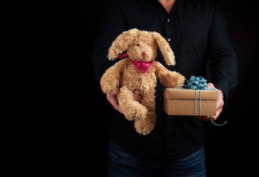 adult man stands on a dark background wearing a black shirt and holds in his hand a brown cardboard box with a gift and a cute brown rabbit toy, happy birthday greetings concept