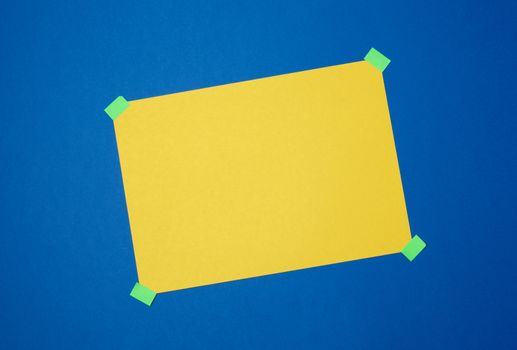 blank yellow sheet of paper stuck with green sticky tape on a blue background, place for text