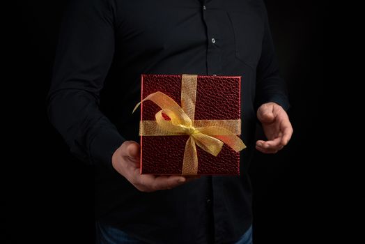 adult man in a black shirt holds a red square box with a knotted golden bow on a dark background, concept of congratulations, surprise