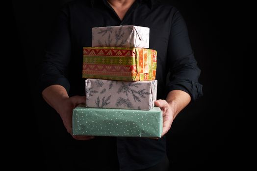 adult man in a black shirt holds in his hands a stack of paper-wrapped gifts on a black background, concept of holiday greetings, surprise and gift