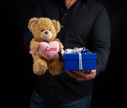 adult man in a black shirt holds a blue square box tied with a white ribbon and brown teddy bear with a heart, concept of congratulations on Valentine's Day on February 14, low key 