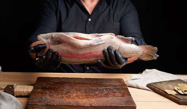 chef in a black shirt and black latex gloves holds a raw carcass of headless salmon fish over a brown wooden cutting board, process of cutting fish