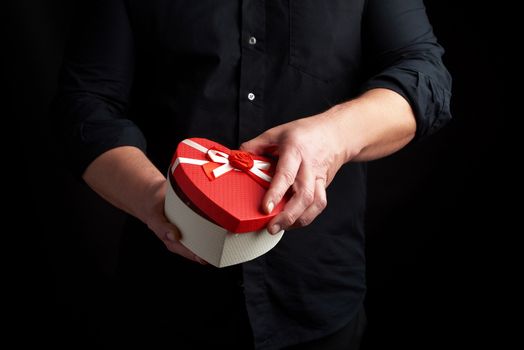 adult man in a black shirt holds a red cardboard box in the form of a heart with a bow on a dark background, concept of holiday poisoning, surprise and a gift