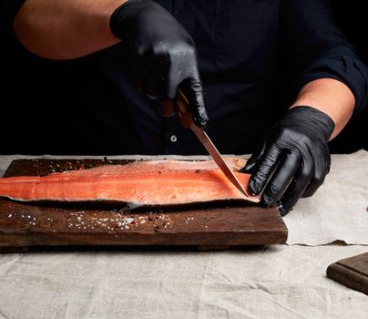 chef in black clothes and black latex gloves cuts fresh salmon filet into pieces, dark key