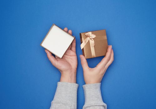 female hand in a gray sweater holds open square cardboard gift box with a bow on a blue background, trend colors, holiday backdrop