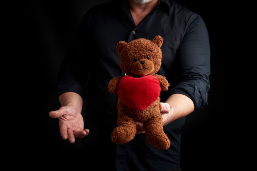 adult man in a black shirt holds a brown teddy bear with a red heart on a dark background, concept of congratulations on the holiday of Valentine's Day on February 14, low key