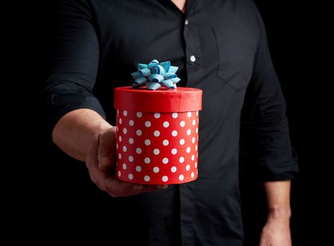 adult man in a black shirt holds in his hand a round red box of polka dots with a blue bow on a black background, concept of a holiday greetings, surprise and gratitude