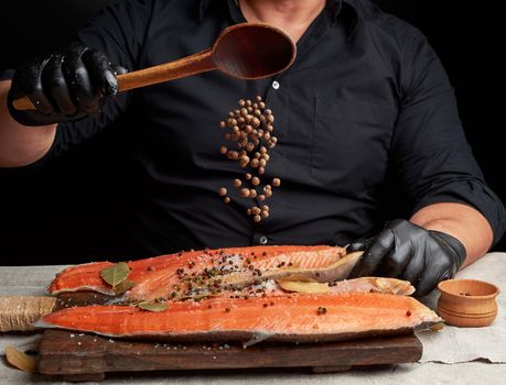 chef in black clothes and black latex gloves pours allspice on a sliced carcass of fresh salmon fish, low key, spices soar in the air