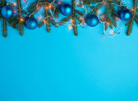 burning multi-colored light bulbs on a garland with a white cable and green twigs, blue balls, blue background, copy space