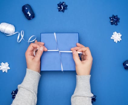 two female hands in a gray sweater hold closed square blue box with a bow on a blue classic background, top view, festive backdrop