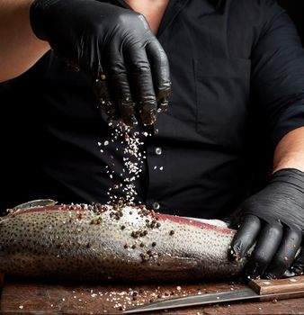 chef in a black shirt and black latex gloves prepares salmon fillet on a wooden cutting board, process of sprinkling with spices and salt, low key