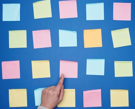 female hand and a lot of empty paper multi-colored square stickers on a blue background, concept of choosing a strategy with the specified finger