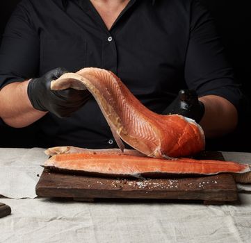 chef in a black shirt and black latex gloves holds a large piece of salmon fish fillet over the table, cooking process, clouse up