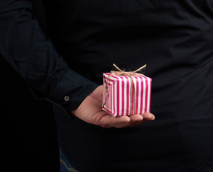 male hand holds a paper-wrapped box with a gift behind his back, man is wearing a black shirt, a concept of surprise