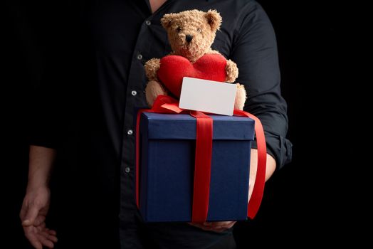 adult man in a black shirt holds a blue square box tied with a red ribbon and sits on top of a brown teddy bear with a heart, concept of congratulations on Valentine's Day on February 14