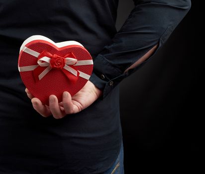 man’s hand holds a cardboard red box with a bow behind his back, a man wearing a black shirt, a concept of surprise