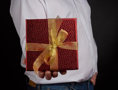 adult man in a white shirt holds a red square box with a knotted golden bow on a dark background, concept of congratulations, surprise