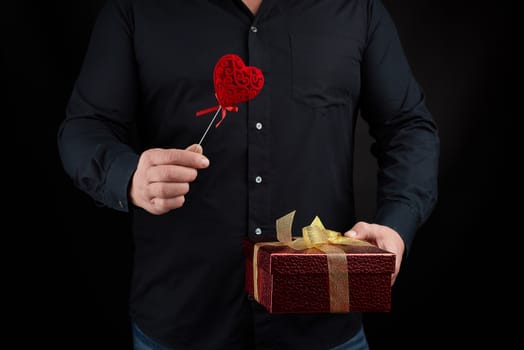 an adult man in a black shirt holds in his hand a red cardboard box tied with a golden bow and a red heart, holiday greetings concept, surprise