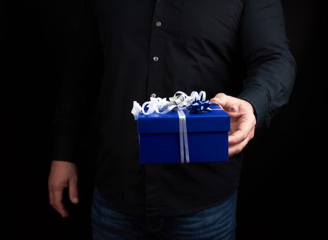 adult man in a black shirt holds a blue square box with a white bow tied on a dark background, concept of congratulations, surprise