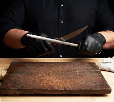 chef in a black shirt and black latex gloves sharpen a kitchen knife on an iron sharpener with a handle above the table, low key