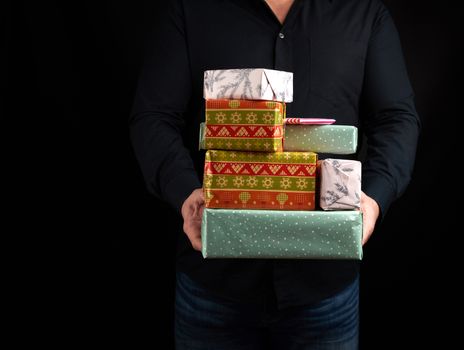 adult man in a black shirt holds in his hands a stack of paper-wrapped gifts on a black background, concept of holiday greetings, surprise and gift