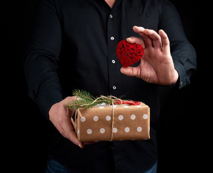 adult man is standing on a dark background wearing a black shirt and holding a wrapped gift in brown paper in his hand, and a red carved decorative heart in his left hand, concept of a holiday greetings