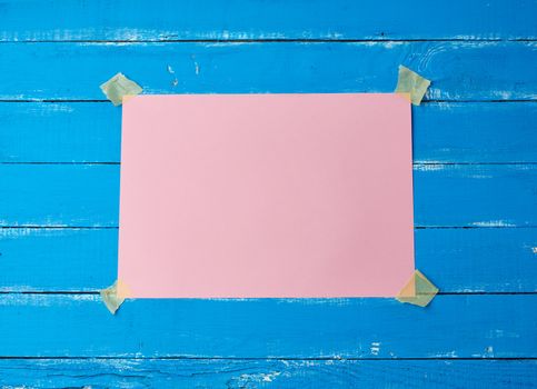 glued with adhesive tape a rectangular empty pink sheet of paper on a blue wooden background, place for an inscription
