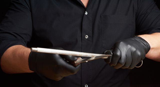 chef in a black shirt and black latex gloves sharpen a kitchen knife on an iron sharpener with a handle above the table, low key, close up