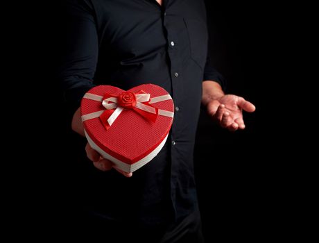 adult man in a black shirt holds a red cardboard box in the form of a heart with a bow on a dark background, concept of holiday greetings, surprise and a gift