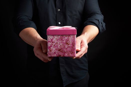 adult man in a black shirt holds a pink square box with a bow tied on a dark background, concept of congratulations, surprise
