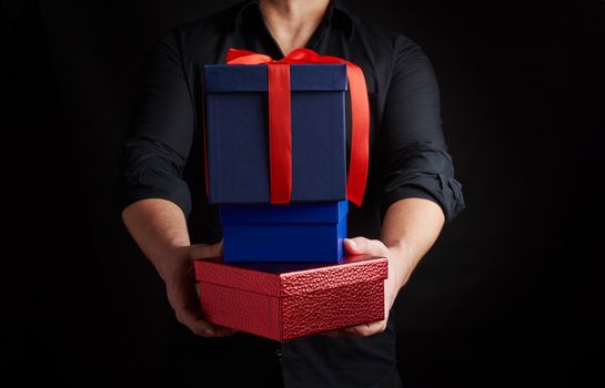 adult man in a black shirt holds in his hands a stack of paper-wrapped gifts on a black background, concept of holiday greetings, surprise and gift