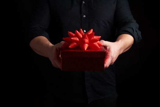 adult man in a black shirt holds a red square box with a knotted bow on a dark background, concept of holiday greetings, surprise