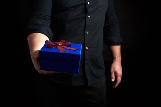 adult man in a black shirt holds a blue square box with a red bow tied on a dark background, concept of holiday greetings, surprise