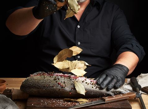 chef in black uniform and black latex gloves pours dry leaves of bay leaf onto cut fresh salmon fillet, seasoning scatters in different directions