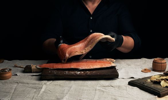 chef in a black shirt and black latex gloves holds a large piece of salmon fish fillet over the table, cooking process, clouse up