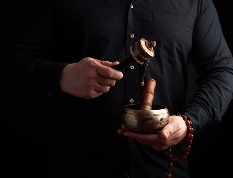 man in a black shirt holds a Tibetan brass singing bowl and a wooden stick and a prayer drum, a ritual of meditation, prayer and immersion in a trance. Alternative treatment