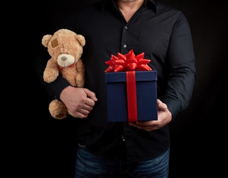adult man in a black shirt holds a blue square box tied with a red ribbon and brown teddy bear, concept of congratulations on Valentine's Day on February 14, low key 