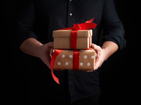 adult man in a black shirt holds a stack of gifts wrapped in brown paper with a red bow on a black background, concept of holiday greetings, surprise and gift