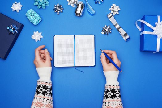 open notebook with empty white sheets and two female hands, blue background with box and decor for gift wrapping, top view