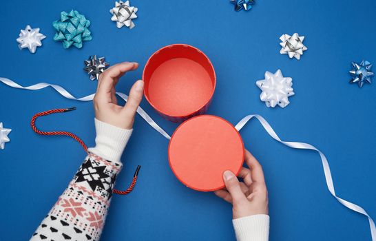 empty open red cardboard box, female hands hold the lid, blue background with decor elements for gift wrapping, top view