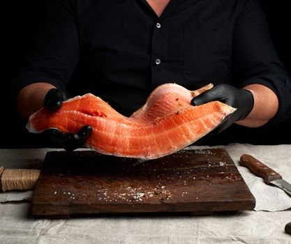 chef in a black shirt and black latex gloves holds a large piece of salmon fish fillet over the table, cooking process, clouse up