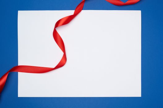 empty white sheet of paper and red silk twisted ribbon on a blue background, place for test, holiday backdrop