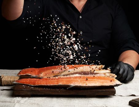chef in a black shirt and black latex gloves prepares salmon fillet on a wooden cutting board, process of sprinkling with spices and salt, low key