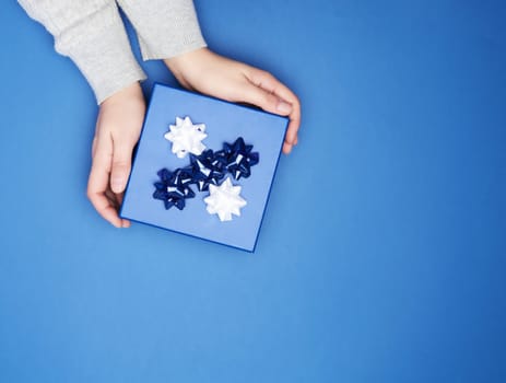 two female hands in a gray sweater hold closed square blue box with a bow on a blue classic background, top view, festive backdrop