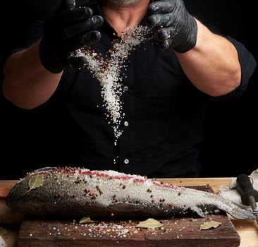 chef in a black shirt and black latex gloves prepares salmon fillet on a wooden cutting board,  process of sprinkling with spices and salt, low key