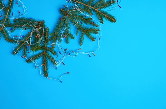 burning multi-colored light bulbs on a garland with a white cable and green twigs of needles, blue background, copy space