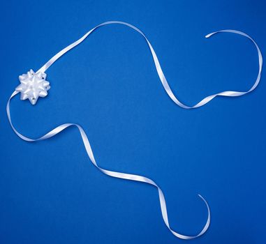 white bow and white silk thin ribbon twisted on a blue background, trendy classic color, top view