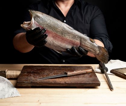 chef in a black shirt and black latex gloves holds a raw carcass of headless salmon fish over a brown wooden cutting board, process of cutting fish